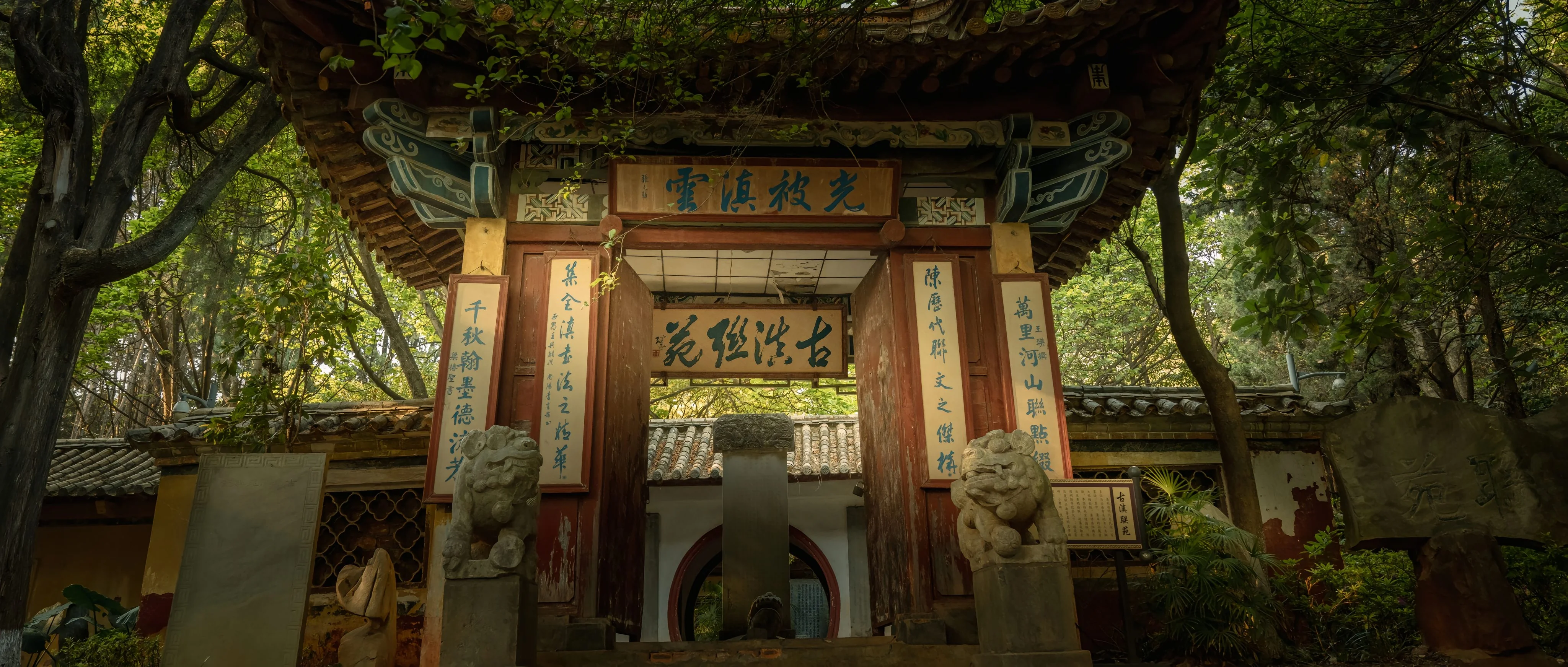 The traditional Chinese entrance gate at a Kunming heritage site, featuring stone lions, blue calligraphy plaques reading "Guang Bei Zhen Yun," and ancient stone steles.
